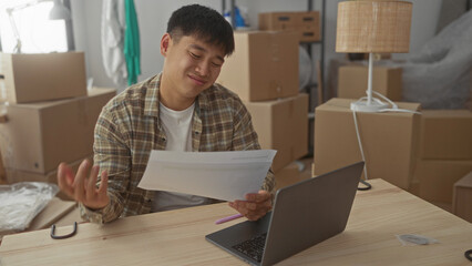 Young man in new home surrounded by boxes working on laptop and documents at wooden table in bright...