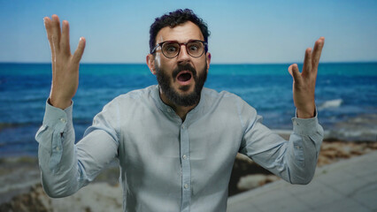 Naklejka premium Hispanic man with beard and glasses expresses surprise on a seaside promenade against a backdrop of blue sea and clear sky.