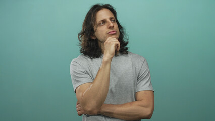 Man with long hair hand on chin in teal studio wearing gray t shirt, arms folded and contemplative...