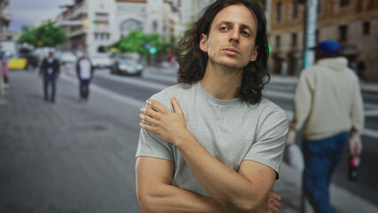 Man with long hair, hand to chin and crossed arms on city street, forearms exposed in resting pose; quiet contemplation.