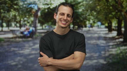 Fototapeta premium Man smiling with arms crossed showing forearms on a sunlit park pathway under green trees and dappled shade; confidence ease.