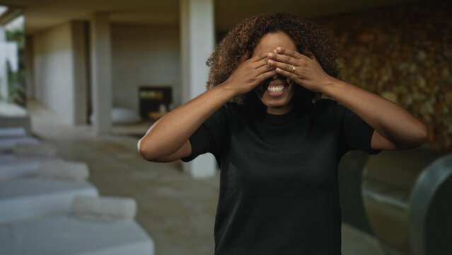Woman smiling and laughing with hands on temples and covering eyes in a spa building with poolside loungers and indoor columns visible; joy relaxation.