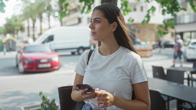 Woman holding smartphone and scrolling at a sidewalk cafe on a busy street during daylight; urban calm.