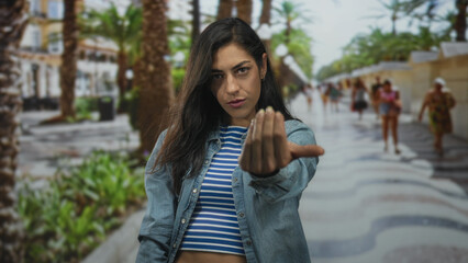 Fototapeta premium Young woman beckons with outstretched hand toward camera on street promenade lined with palm trees and buildings; assertive invitation.