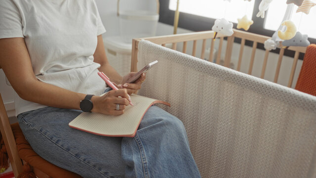 Woman writing with pen and holding smartphone by a crib in a nursery, hands on notebook across denim lap, seated indoors; calm planning motherhood.