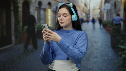 Fototapeta premium Woman in blue sweater with headphones uses smartphone on cobblestone street in beautiful old city, surrounded by historic architecture.