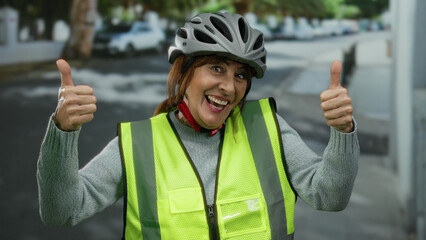 Hispanic woman wearing a bicycle helmet and safety vest gives thumbs up on a city street, showcasing vibrant positivity and outdoor enthusiasm.