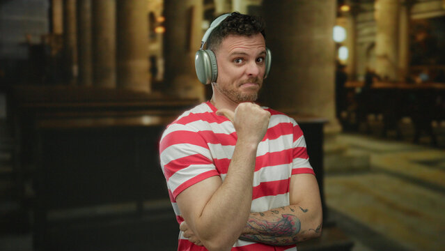 Young man with tattoos wearing headphones and striped shirt points thumb indoors in church setting, looking relaxed and thoughtful.