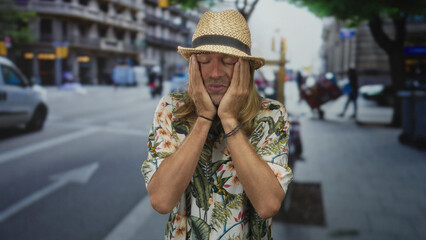 Man covering face with hands wearing floral shirt amid sunny busy urban street traffic; embarrassment.
