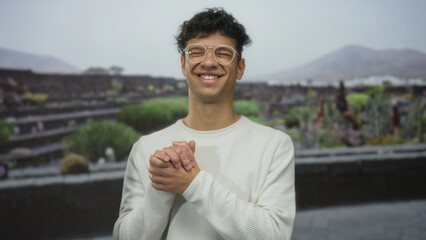Fototapeta premium Man clasps hands and smiles through clear glasses in front of weathered stone ruins and a building wall; calmness.