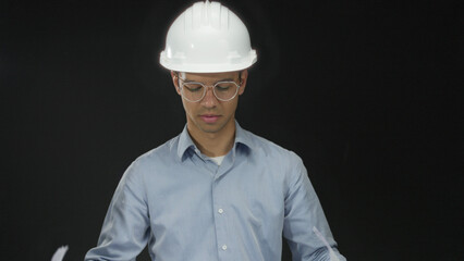 Man engineer wearing white hardhat and clear glasses reading large blueprint sheet in studio; concentration.