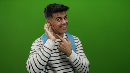 Young man in striped shirt smiling with hand to ear over isolated green background, depicting a...