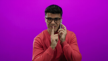 Handsome man wearing glasses in red sweater talks on phone against vibrant pink background making gesture for silence