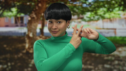 Young chinese woman in green shirt makes hashtag gesture outdoors in sunny park setting with trees and blurred buildings in background highlighting casual social interaction.