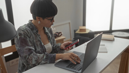 Woman working on laptop while checking smartphone in modern living room setting at home.