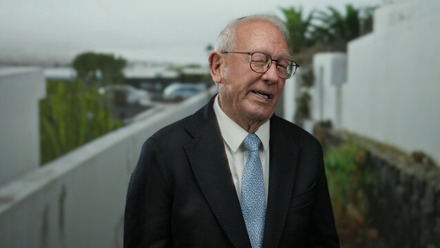 Senior man in a business suit winks while standing outdoors on a street, conveying confidence and charm with a light-hearted expression. - Powered by Adobe