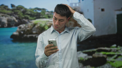 Fototapeta premium Young hispanic man scratches head holding smartphone beside a white building by the turquoise seaside; confusion.