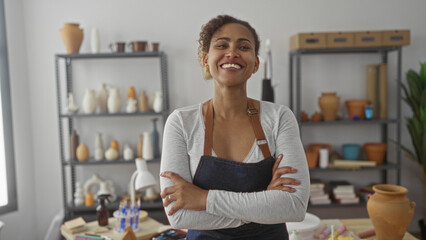 Woman wearing apron with arms crossed and smiling among ceramic pots on metal shelves, table of clay tools and a vase in studio; craft pride.