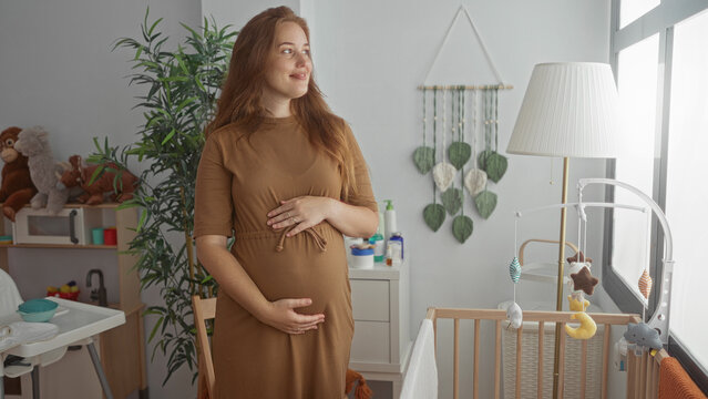 Pregnant woman gently touches belly beside wooden crib adorned with baby mobile in bright sunlit nursery; serene anticipation.