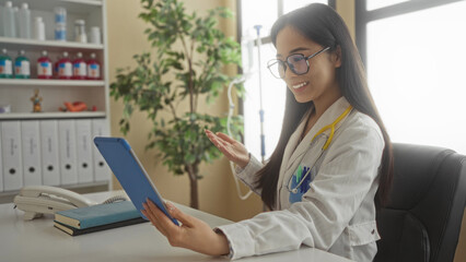Doctor holding tablet during video call in hospital room, smiling warmly, surrounded by medical...