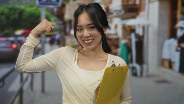 Woman flexing arm with a smile holds yellow clipboard on a busy street showcasing confidence and strength in an outdoor urban setting with a vibrant, lively backdrop.