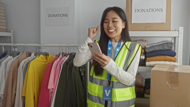 Young woman volunteer in reflective vest uses smartphone in charity center surrounded by donations and clothing racks indoors.
