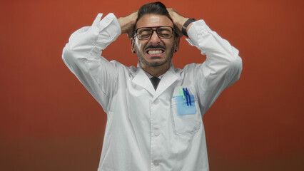 Young hispanic scientist in lab coat grips head with both hands and clenches teeth in orange studio; stress.