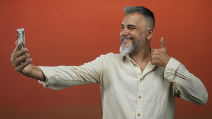 Man middle age taking a selfie with smartphone and thumbs up pose in orange studio set; confidence connection.