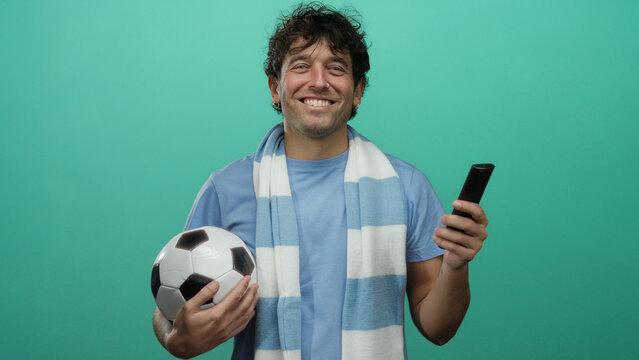 Hispanic man holding football and remote on green background, smiling enthusiastically, capturing the essence of sports fandom and leisure.