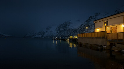 A winter night fjord in Norway. The mountain slopes are covered with snow. A small building is illuminated at night.