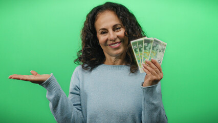 Middle-aged woman holding israeli shekels smiling against a vibrant green background, showcasing...