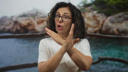 Middle age hispanic woman crosses arms in rejection gesture on rocky beach; denial refusal...