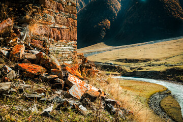 Close-up of ancient stone wall with vivid orange lichen in Truso Valley, Georgia. Autumn sunlight highlights textures, with a winding river and mountains in the background