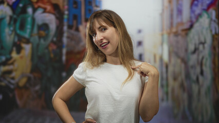 Naklejka premium Woman points finger to chest while smiling on graffiti covered street in a white t shirt and relaxed pose; confidence.