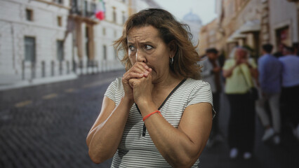 Hispanic middleaged woman wearing striped top clasps bare hands to chin on cobblestone street with historic building facades in warm daylight; thinking doubt concern.