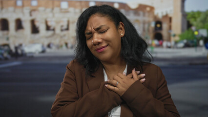 Fototapeta premium Woman clutching her chest with closed eyes and furrowed brow in front of roman coliseum outdoors during daylight; pain.