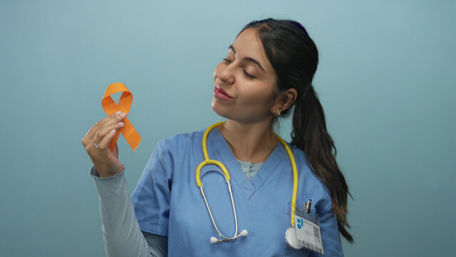 Woman doctor in blue uniform holding orange ribbon with confident smile against isolated blue background representing health awareness.