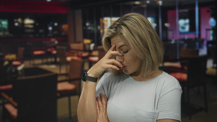 Blonde woman holds cheek and gazes upward in restaurant interior with blurred tables; thoughtful reflection.