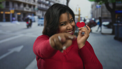 Fototapeta premium Latin woman in red holds a heart-shaped paper, smiling confidently on a bustling street city backdrop, embodying love and positivity outdoors.
