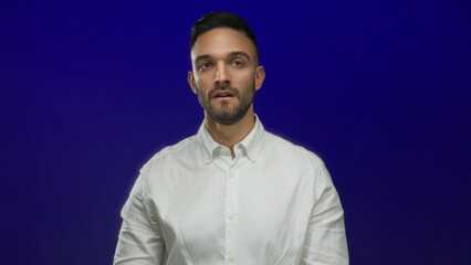 Obraz premium Tired young hispanic man in a white shirt poses thoughtfully against a solid blue background, conveying a sense of fatigue and introspection.