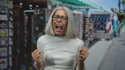 Obraz premium Middle aged woman with clenched fists shouting in a busy street souvenir market aisle, grey hair and glasses, wearing white top; frustration outburst.
