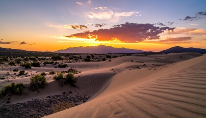 Desert landscape at sunset, featuring undulating sand dunes in the foreground, shrubs, and distant mountains under a colorful, cloudy sky