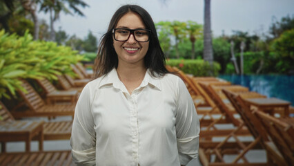 Woman smiling with glasses, face visible, standing by pool lounge chairs at a resort hotel building; serenity.
