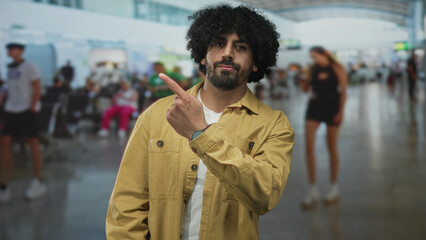 Naklejka premium Man with curly hair pointing finger upward while looking up in busy airport terminal; curiosity discovery.