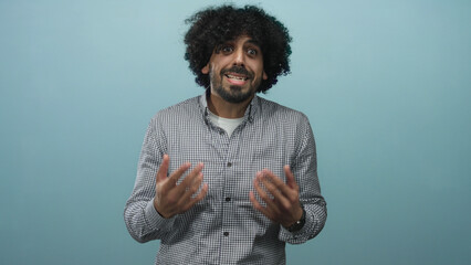 Man with curly hair and beard shrugging with open palms in light blue studio wearing check shirt...