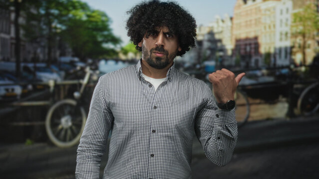 Man with curly hair and beard points thumb over shoulder on street in amsterdam beside a canal and parked bicycles while wearing a checked shirt; skepticism.