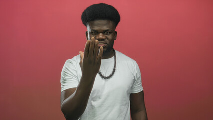 Man in white t shirt extends hand toward camera in studio with red wall and beaded necklace,...