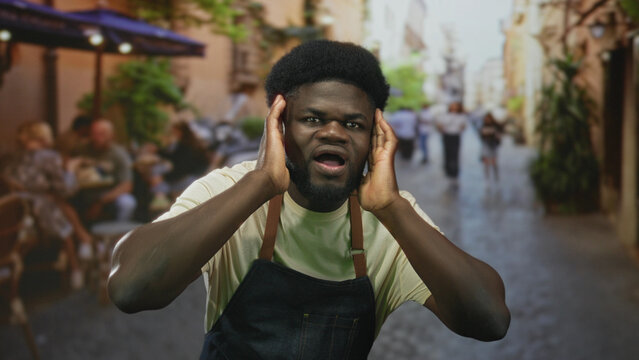 Young african american man cupping hands to shout on street terrace restaurant wearing denim apron and smiling; friendly welcome. - Powered by Adobe