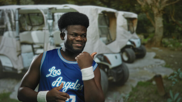 Man with thumbs up and finger pointing gesture in forest near parked golfcarts wearing blue los angeles jersey; playful confidence.