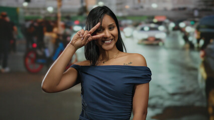Fototapeta premium Woman smiling on a bustling street at night, making a peace sign while wearing a blue dress, capturing a joyful moment in dynamic urban surroundings.
